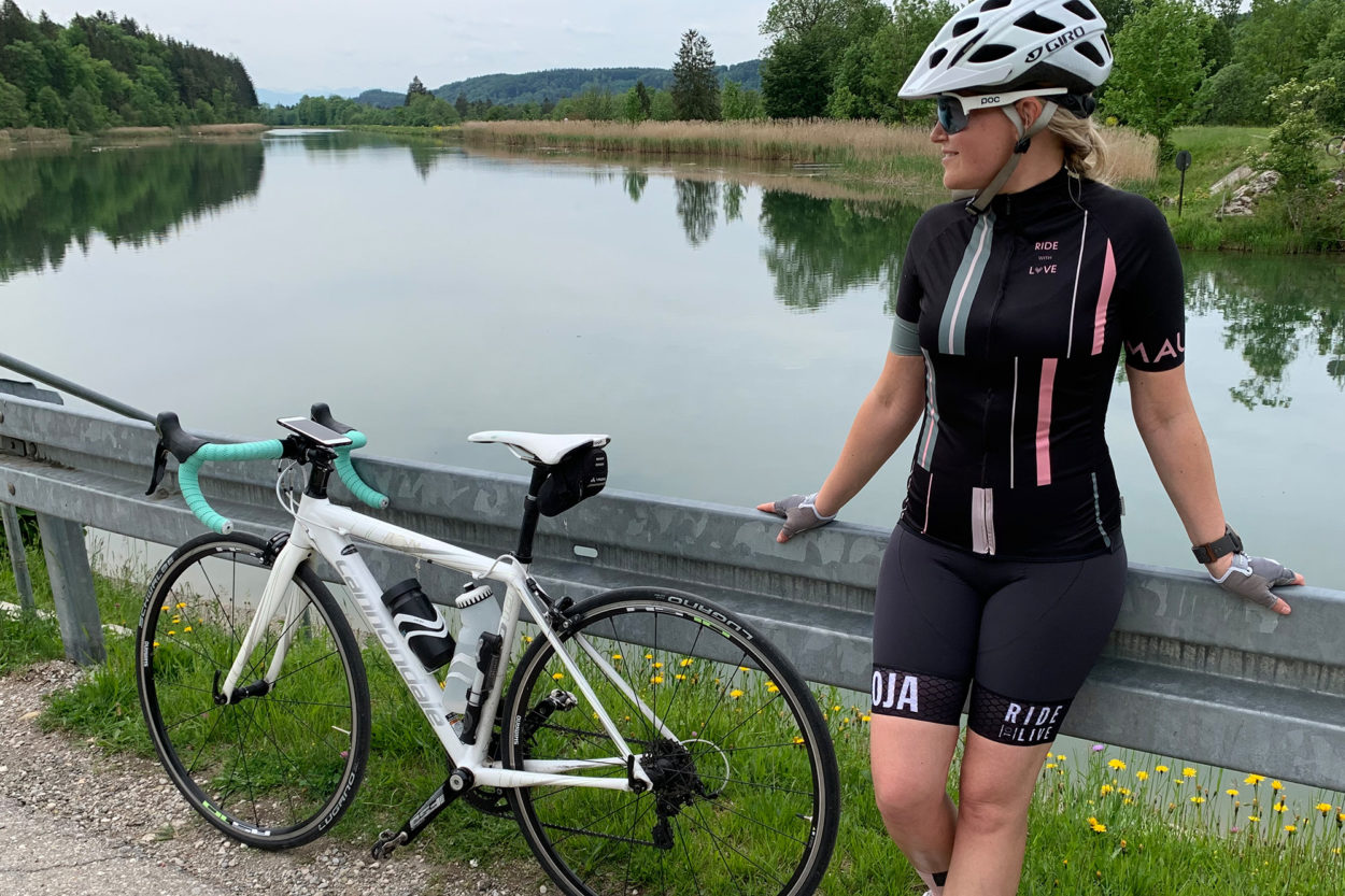 Frau steht neben ihrem Rennrad an eine Leitplanke gelehnt. Im Hintergrund ein Fluss.